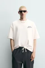 Camiseta blanca de corte relajado, con cuello redondo, manga corta y texto estampado en contraste en el pecho.