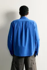Camisa de corte relajado confeccionada en tejido de algodón de color azul, con cuello abotonado, manga larga, bolsillo frontal de parche y cierre de botones.