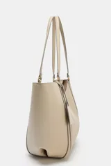 Bolso tipo shopper de hombro en color beige, con diseño estructurado, doble asa y detalle de pespuntes verticales en el cuerpo. Incluye un colgante decorativo tipo etiqueta y cierre imantado.