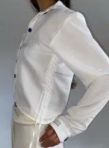 Camisa blanca de corte cropped con ajuste mediante cordones laterales y botones en contraste de color azul oscuro. Presenta cuello clásico, mangas largas con puños y diseño ligero.
