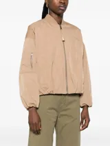 Campera bomber color beige, corte regular, con cierre frontal con cremallera visible, cuello acanalado, puños y bajo elásticos fruncidos, hombro caído, bolsillos de ribete verticales y bolsillo utilitario con cremallera en la manga.