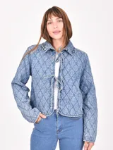 Sobrecamisa acolchada de denim azul claro con patrón de rombos. Presenta cuello con detalle de encaje o puntilla y cierre frontal mediante lazos anudados.