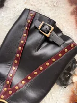 Bota alta de cuero negro con taco cuadrado de efecto madera, puntera redondeada y detalles de tiras en tono bordó con tachas metálicas doradas. Cuenta con una argolla metálica en el empeine y una hebilla de ajuste en la parte superior de la caña.