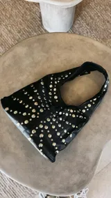 Cartera de hombro de cuero sintético negro, con diseño hobo y decoración de tachas metálicas plateadas dispuestas en patrón radiante.