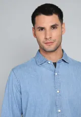 Camisa de jean azul con botones blancos, mangas largas con puños doblados y corte clásico.