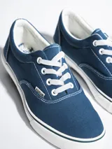 Championes de lona color azul grisáceo, estilo skate o casual, con cordones blancos y suela de goma blanca con una línea fina azul en el borde.