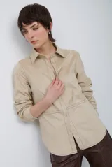 Camisa de gamuza sintética en tono beige, con cuello clásico, cierre frontal de botones y costuras decorativas visibles. Presenta mangas largas con puños abotonados y un corte recto con ruedo redondeado.