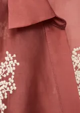 Camisa de manga larga en color terracota con diseño semitransparente. Presenta cuello camisero, cierre frontal con botones y bordados florales en color blanco distribuidos por toda la prenda.