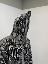 Buzo con cierre frontal completo, confeccionado en tejido de punto grueso de algodón. Presenta un diseño estampado integral con motivos gráficos y tipografía estilo gótico en tonos gris y negro, capucha integrada, puños y ruedo acanalados en color negro sólido.