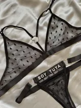 Conjunto de lencería de dos piezas en tul negro con estampado de corazones floca, compuesto por un bralette con tiras ajustables y argolla en forma de corazón, y una tanga con tiras regulables, argollas metálicas y detalle de pedrería con la inscripción 'Soy Tuya'.