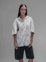 Camisa de manga corta color beige con cuello clásico y cierre de botones. Presenta un diseño de bordado calado con motivos florales y geométricos.