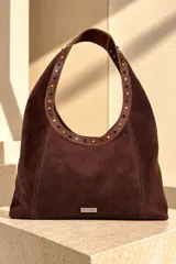 Cartera tipo hobo de gamuza color marrón chocolate, con tira de cuero decorada con tachas metálicas doradas y negras. Incluye placa metálica con el logo de la marca en la base.