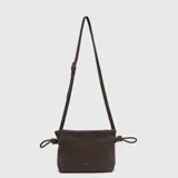 Cartera tipo bandolera de color marrón oscuro, con textura granulada, cierre superior y detalles de nudos en los laterales. Incluye una correa larga ajustable y removible.