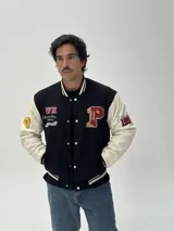 Campera estilo varsity con cuerpo negro y mangas blancas. Presenta parches bordados en el pecho y las mangas, cierre frontal con botones a presión y puños, cuello y ruedo acanalados con rayas blancas y negras.