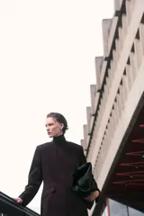 Vestido corto de sarga color marrón oscuro, con escote redondo, manga larga y cintura entallada con pinzas. Presenta bolsillos ribeteados con solapa en la parte delantera.