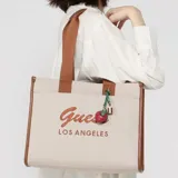 Cartera tipo tote bag Guess modelo Jackie, confeccionada en algodón color beige con detalles y asas en marrón. Cuenta con el logotipo de la marca bordado en el frente y un charm colgante en forma de cereza con un pequeño candado dorado.