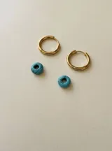 Aros de acero quirúrgico dorado en forma de argolla con dijes desmontables de piedra natural turquesa con vetas negras, en forma de doña.
