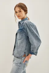 Camisa de jean de corte clásico con efecto lavado, cuello camisero, cierre frontal con botones metálicos y bolsillos frontales con solapa. Presenta mangas largas con diseño abullonado en los hombros y puños abotonados.