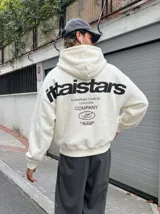 Hoodie de color crema con capucha, manga larga y diseño holgado. Presenta un estampado gráfico en la espalda con el texto 'ittai stars' y detalles de marca en color negro.