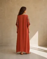 Vestido largo color terracota de lino, con diseño asimétrico que incluye un panel lateral estampado con grandes flores oscuras, escote redondo, mangas 3/4 con trabilla y botón para arremangar, y un amplio bolsillo frontal aplicado en el pecho con terminaciones deshilachadas.