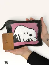Sobre de cuero negro con intervención artística pintada a mano que representa una figura blanca sobre fondo rosado. Incluye cierre superior y correa de muñeca.