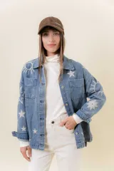 Camisa de denim de lavado medio, con calce relajado y ligeramente oversize. Presenta bordados o estampados de inspiración celestial (soles, lunas y estrellas) en color claro en la parte delantera y mangas. Tiene cuello, bolsillos delanteros con tapa y cierre frontal con botones.