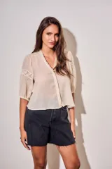 Blusa de color arena con cuello en V, mangas tres cuartos abullonadas y detalles de encaje bordado en hombros y mangas. Presenta un diseño ligero y delicado con cierre frontal.