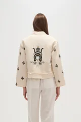 Campera estilo bomber color beige claro, con cuello redondo, cierre frontal y detalles de volados en los bordes. Presenta bordados florales en negro y blanco en la zona del pecho y bordados de tréboles negros en las mangas.