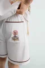Bermuda de corte relajado confeccionada en algodón, color blanco con vivo en contraste color marrón en el ruedo. Presenta cintura elástica con cordón ajustable y un bordado decorativo en la pierna derecha que incluye una palmera y un gallo.