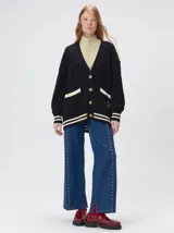 Cardigan de punto trenzado en color negro con detalles en color crema en los bolsillos, puños y ruedo. Presenta un diseño oversize, cierre frontal con botones y bordado decorativo en uno de los bolsillos delanteros.