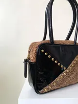 Cartera rígida negra con detalles en rojo y tachas plateadas. Tiene herrajes y cierre metálico plateado marca YKK, asa fina para llevar al hombro o en la mano y correa larga extra regulable. El interior es forrado, tiene un bolsillo con cierre enmarcado en cuero, y un segundo bolsillo plaqué.