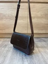 Cartera bandolera de cuero vacuno color bordó, con solapa frontal de textura símil cocodrilo y detalles de costuras pespunteadas en blanco. Cuenta con correa ajustable y cierre superior.