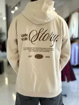 Canguro oversize de color beige con capucha y bolsillo tipo canguro al frente. En la espalda cuenta con un gran estampado tipográfico en marrón con la frase "Own Your Story", texto descriptivo y un logotipo ovalado con las letras "EST 2021".