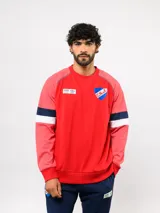 Buzo de piqué color rojo con mangas raglán en un tono de rojo más claro. Presenta franjas horizontales azul marino y blancas en las mangas. Incluye el escudo del Club Nacional de Football y el logo de Umbro en el pecho.
