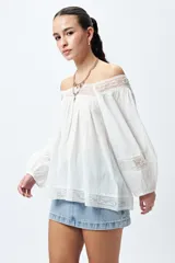 Blusa blanca de algodón con escote off-shoulder, mangas largas con puños elásticos y detalles de bordado y puntillas en escote y mangas.