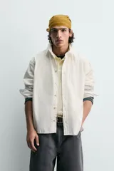 Camisa blanca de algodón con corte relajado, cuello solapa, manga larga con puño abotonado y bolsillo de plastrón en el pecho. Presenta bajo asimétrico con aberturas laterales y cierre frontal de botonadura.