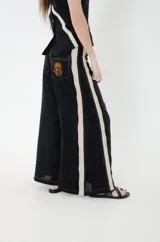 Pantalón de lino negro con franjas laterales en tono beige y pespuntes en hilo color óxido. Presenta corte amplio de piernas anchas, cintura media y un bolsillo trasero con bordado de una rosa en hilo contrastante.