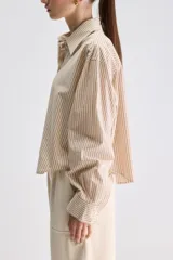 Camisa de calce relajado confeccionada en algodón, con diseño de rayas verticales en tonos beige y blanco. Presenta cuello clásico, cierre frontal con botones, puños abotonados y largo ligeramente cropped.