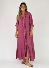 Vestido largo de lino color tostado, con diseño camisero, botonadura frontal completa con detalles de volados a lo largo de la botonadura, mangas largas arremangables y aberturas laterales en el bajo.