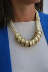 Collar de diseño artesanal compuesto por múltiples tiras de cuero en color crudo y piezas cilíndricas de metal dorado con textura martillada.