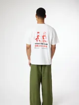 Camiseta blanca de algodón orgánico, con corte oversize, cuello redondo y manguita corta. Presenta un pequeño estampado rojo en el lateral superior izquierdo con la frase 'Go Out And Slide' y caracteres en japonés junto a una ilustración de dos personas cargando una tabla de surf.