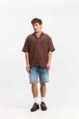 Camisa de manga corta color beige confeccionada en muselina de algodón, con cuello abierto, cierre frontal de botones y un bolsillo aplicado en el pecho.