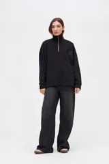 Buzo oversize de felpa con friza, color negro, con cuello alto y cierre frontal parcial metálico. Presenta bolsillos laterales, tajos en los costados y un diseño con espalda más larga. Incluye un pequeño logo bordado en el pecho.