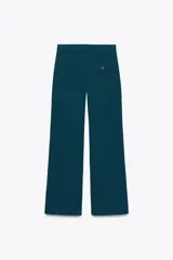 Pantalón de vestir marrón oscuro, tiro alto, con pernera ancha y pinzas frontales. Presenta un bolsillo con tapeta en la parte superior delantera.