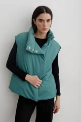 Chaleco puffer de cuello alto con cierre frontal oculto y botones a presión metálicos visibles en la solapa superior. Diseño holgado y acolchado en color verde azulado.