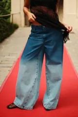 Jean de tiro medio con corte oversize y botamanga ancha. Presenta un diseño de lavado degradado que va de un azul intenso en la cintura a un tono claro en la parte inferior, con efecto gastado. Incluye avíos metálicos y badana de PU.