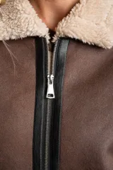 Campera tipo gamulán de corte oversize, color marrón con interior y cuello de corderito sintético en tono crema. Presenta detalles de ribetes en cuero sintético negro a lo largo de la prenda, cierre frontal metálico y puños tejidos.