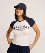 Remera ajustada al cuerpo, estilo raglán, con cuerpo blanco y mangas cortas azul marino. Presenta un estampado en el pecho con el texto "BARCELONA EUROPE TOUR EST. 1992" en azul marino.