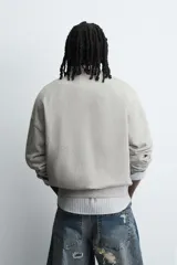Sudadera gris de corte boxy fit, con cuello redondo y manga larga. Presenta un estampado frontal con texto en azul con efecto desgastado y acabados en rib en puños y ruedo.