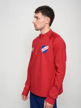 Buzo deportivo de polytraining color rojo, con medio cierre frontal y cuello alto. Presenta el logo de Umbro en el pecho izquierdo y el escudo del Club Nacional de Football en el pecho derecho.